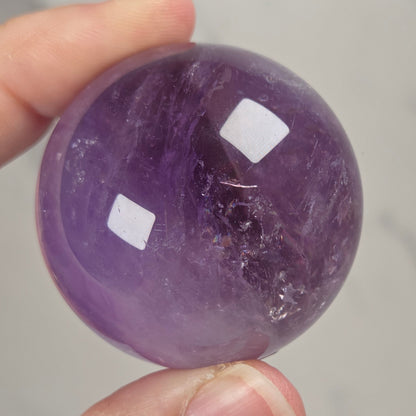 A hand holding a polished purple amethyst crystal sphere against a light gray marble background.
