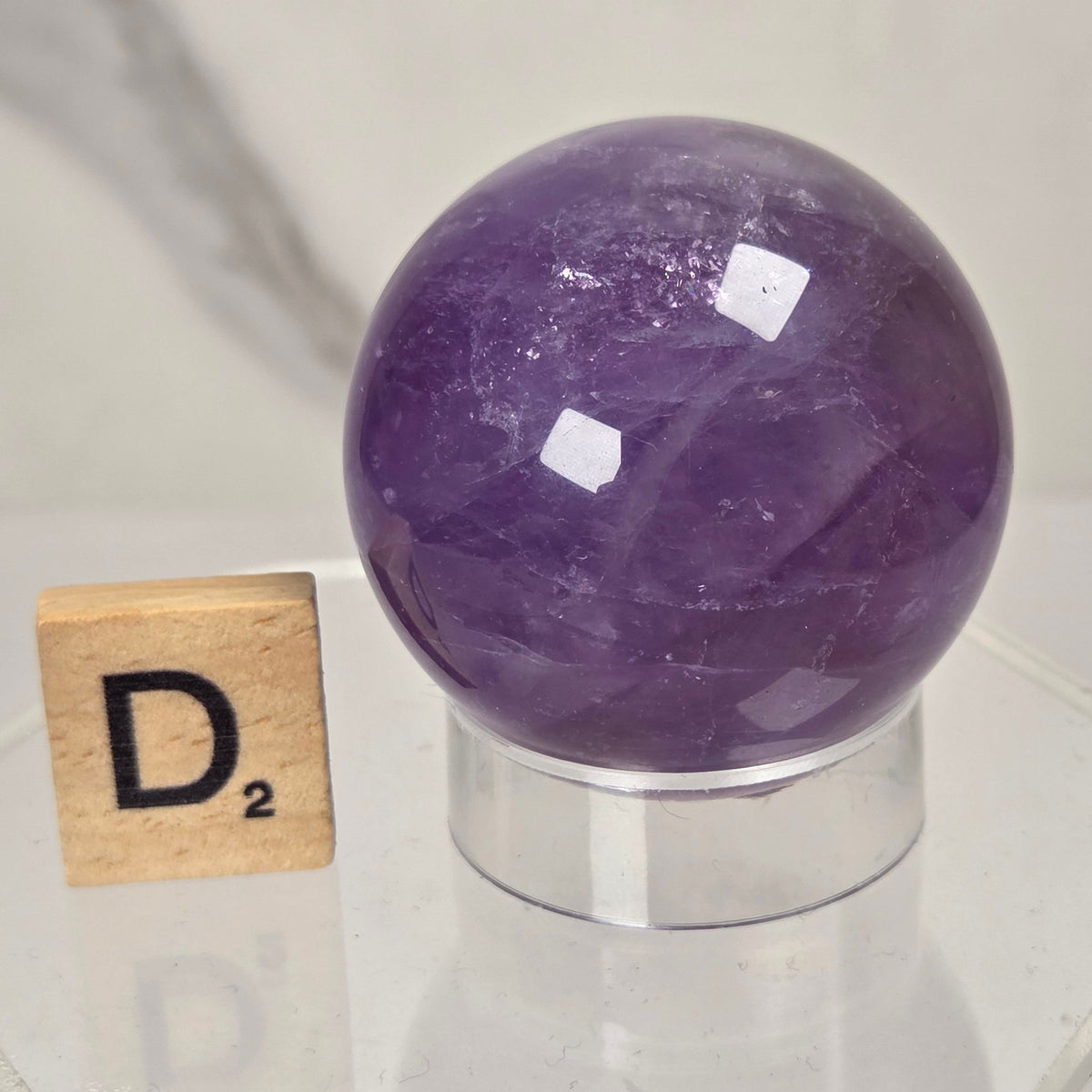 A polished purple amethyst crystal sphere displayed on a clear acrylic stand next to a wooden Scrabble tile showing the letter D2, set against a light gray marble background.