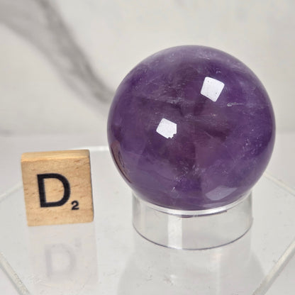 A polished purple crystal sphere displayed on a clear acrylic stand next to a wooden Scrabble tile showing the letter D, set against a soft-focus marble background.