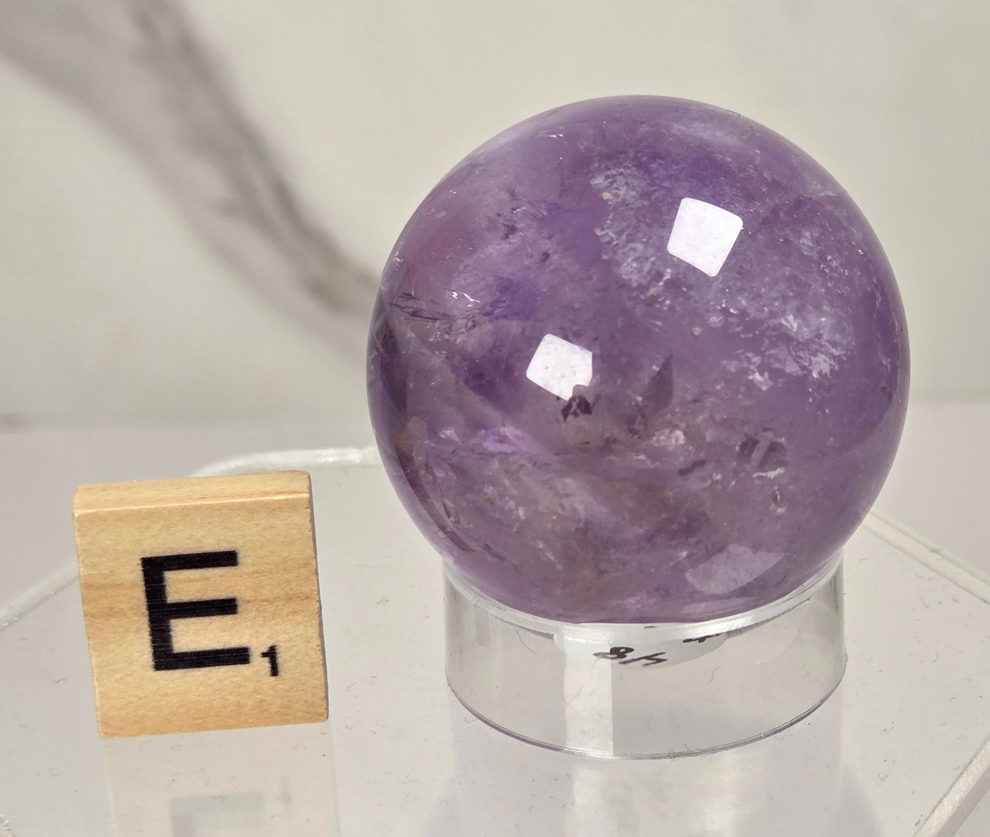 A polished purple amethyst crystal sphere on a clear acrylic stand, shown next to a wooden Scrabble tile with the letter E on a light marble surface.