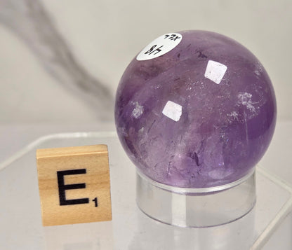 A polished purple amethyst crystal sphere on a clear acrylic stand, next to a wooden Scrabble tile showing the letter E, against a light marble-patterned background.
