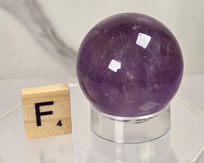 A polished purple crystal sphere on a clear acrylic stand, shown next to a small wooden Scrabble tile with the letter F on a light marble-patterned background.