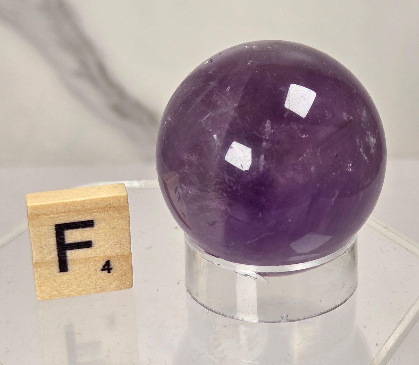 A polished purple amethyst crystal sphere displayed on a clear acrylic stand, with a small wooden Scrabble tile showing the letter F next to it on a reflective surface.