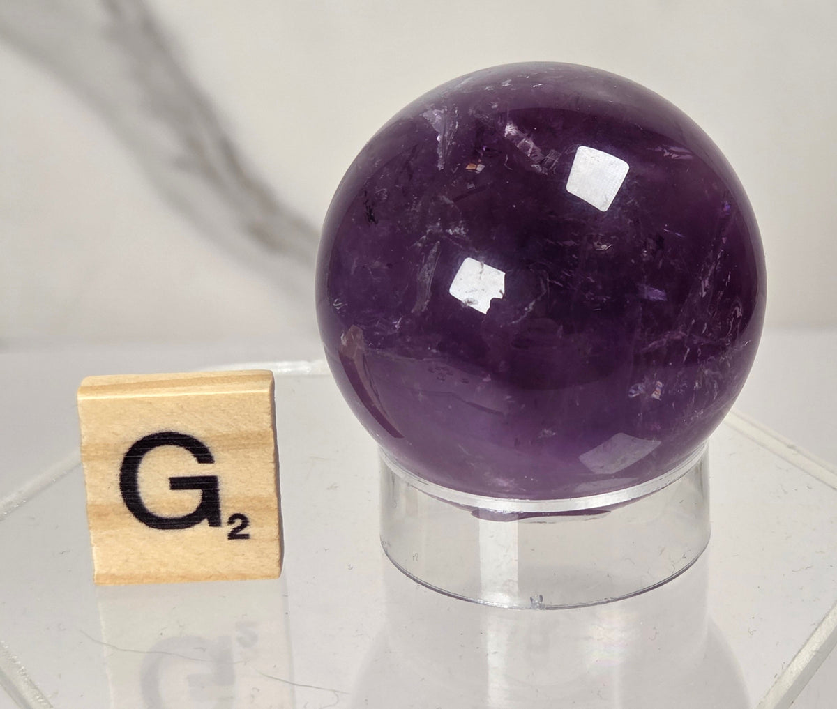 A polished purple amethyst crystal sphere displayed on a clear acrylic stand, with a small wooden Scrabble tile showing 'G2' placed beside it on a reflective white surface.