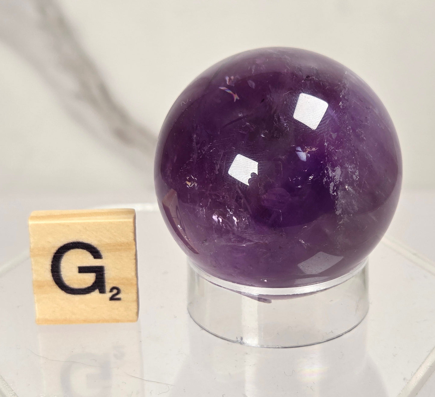A polished purple amethyst crystal sphere displayed on a clear acrylic stand next to a wooden Scrabble tile showing the letter G and value 2, set against a light marble background.
