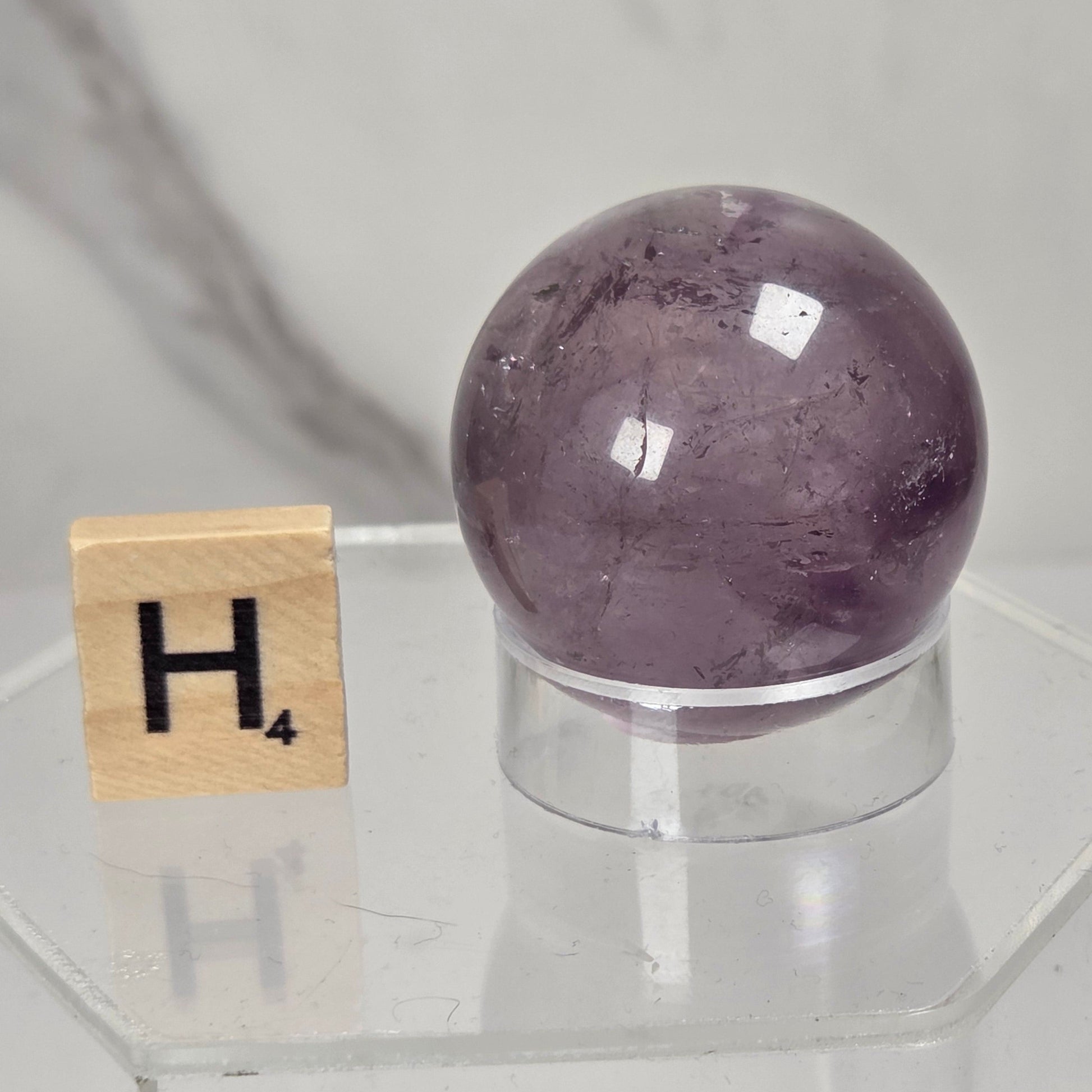 A polished purple amethyst crystal sphere displayed on a clear acrylic stand next to a wooden Scrabble letter tile showing the letter 'H'.