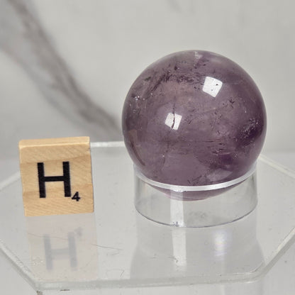 A polished purple amethyst crystal sphere displayed on a clear acrylic stand next to a wooden Scrabble letter tile showing the letter 'H'.