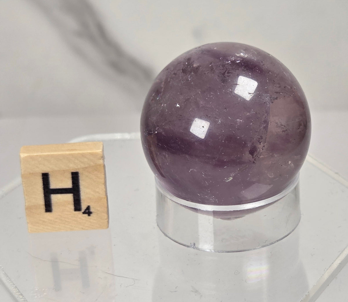 A polished purple amethyst crystal sphere displayed on a clear acrylic stand next to a wooden Scrabble tile showing the letter H, set against a light gray marble background.