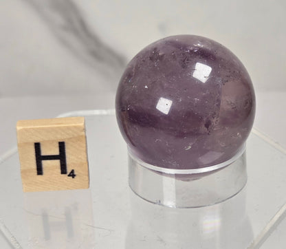 A polished purple amethyst crystal sphere displayed on a clear acrylic stand next to a wooden Scrabble tile showing the letter H, set against a light gray marble background.