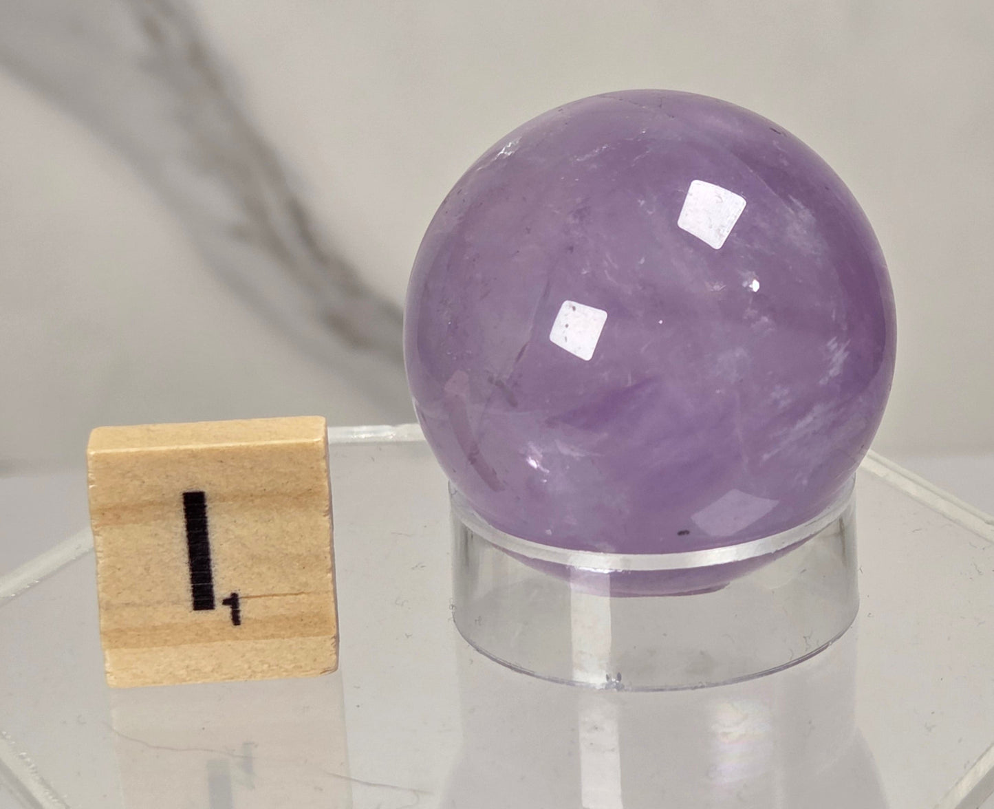 A polished purple amethyst crystal sphere on a clear acrylic display stand, shown next to a small wooden block with the number 1, against a light marble-patterned background.