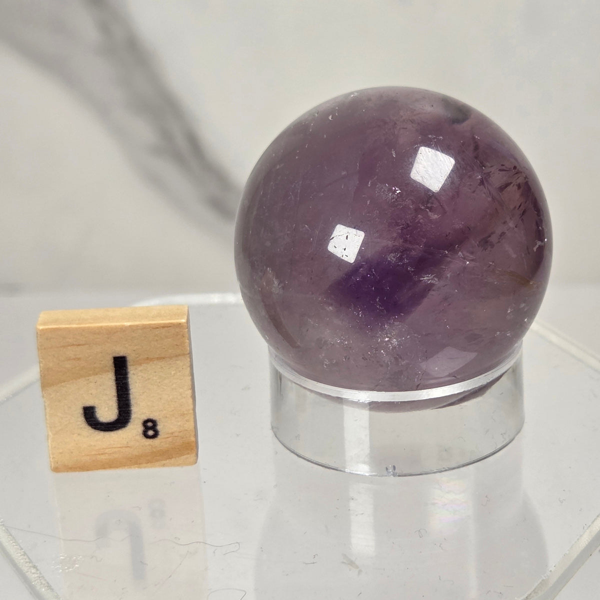 A polished purple amethyst crystal sphere on a clear acrylic stand, shown next to a wooden Scrabble tile with the letter J and 8, against a light marble background.