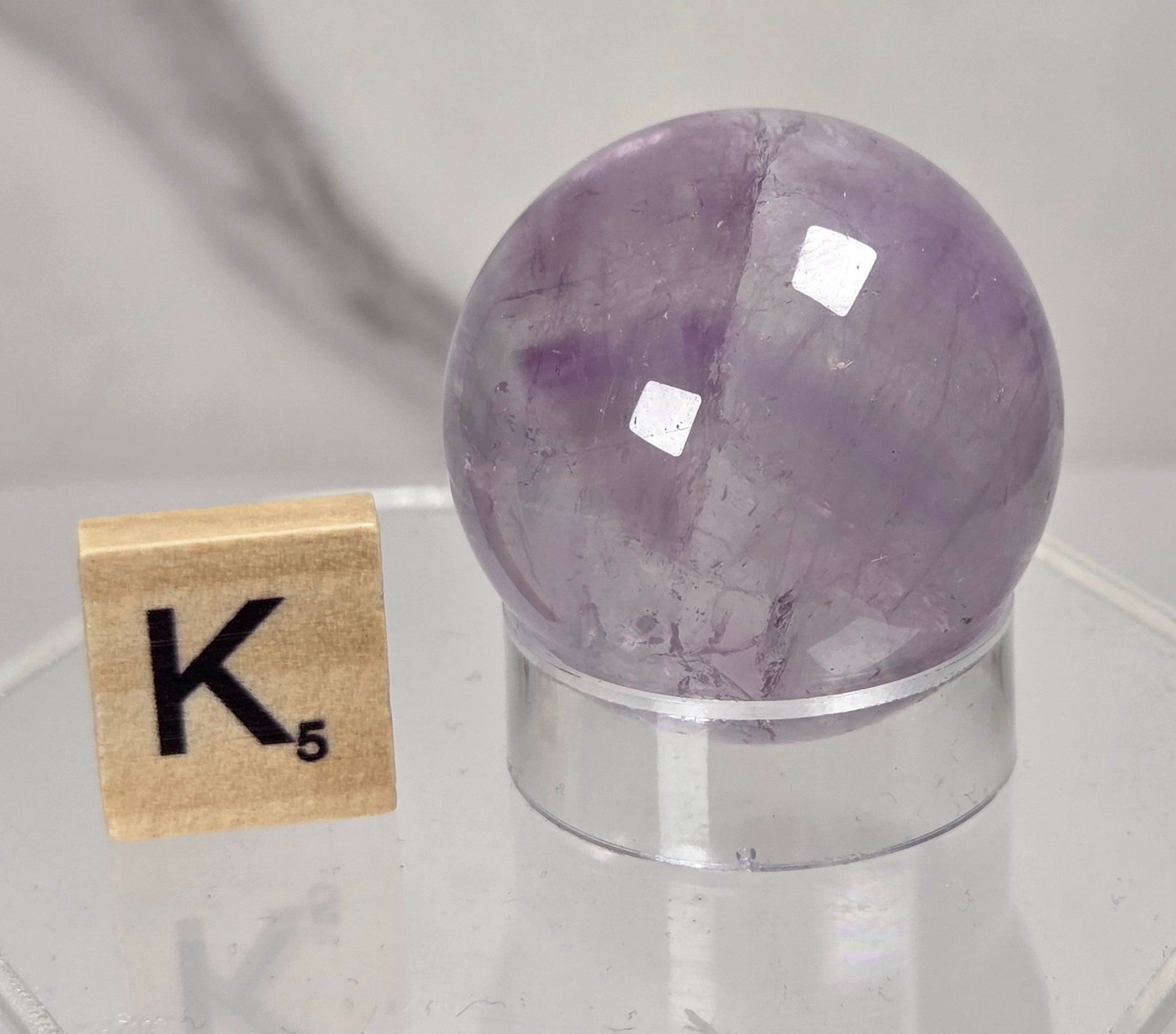 A polished purple amethyst crystal sphere on a clear acrylic stand, shown next to a wooden Scrabble tile spelling K on a light gray marble-patterned background.