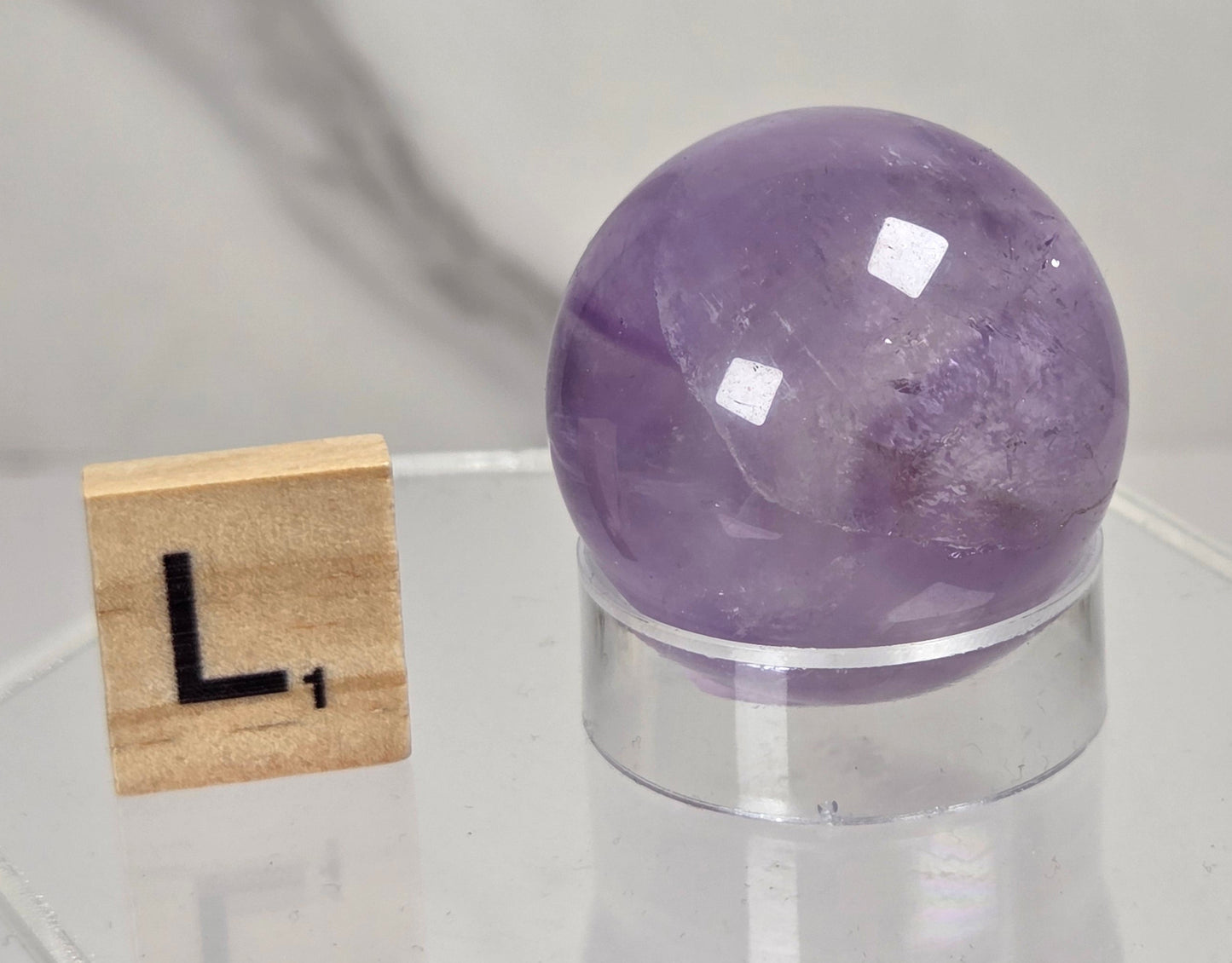 A polished purple amethyst crystal sphere on a clear acrylic stand, displayed next to a wooden Scrabble tile on a clear acrylic hexagonal base against a light gray marble-patterned background.