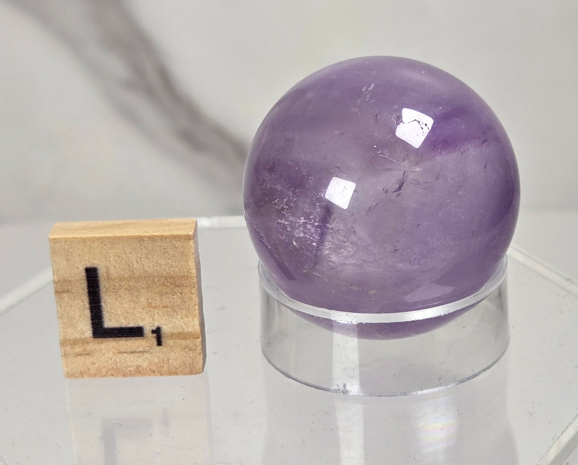 A polished purple amethyst crystal sphere displayed on a clear acrylic stand next to a wooden Scrabble tile showing the letter 'L', set against a light gray marble-patterned background.