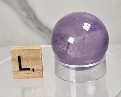 A polished purple amethyst crystal sphere displayed on a clear acrylic stand next to a wooden Scrabble tile showing the letter 'L', set against a light gray marble-patterned background.