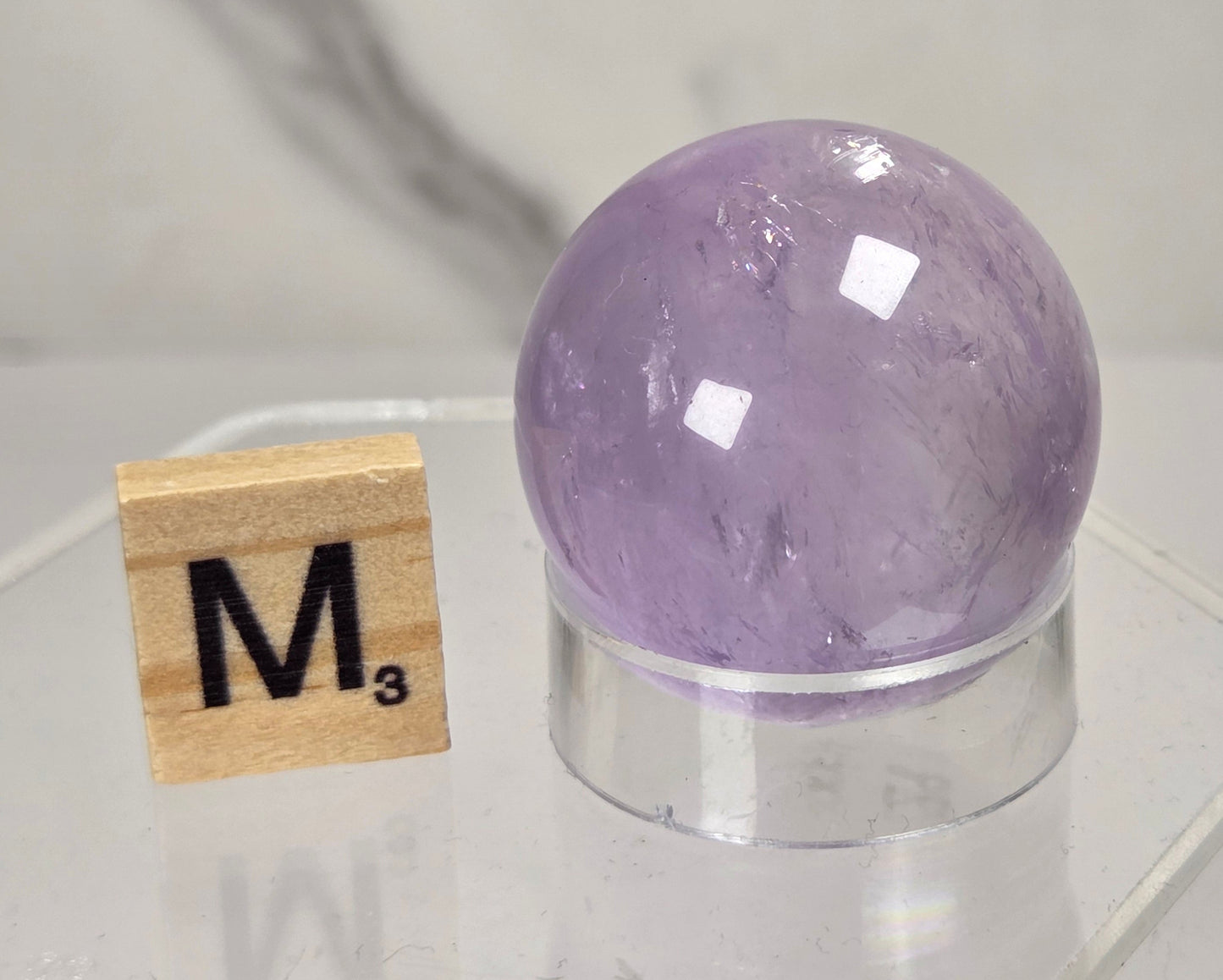 A polished purple crystal sphere displayed on a clear acrylic stand next to a wooden Scrabble tile showing the letter 'M' and the count 'M3', set against a light gray marble-patterned background.