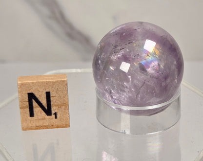 A polished purple crystal sphere resting on a clear acrylic stand, displayed next to a wooden Scrabble tile showing the letter 'N' on a light gray marble-patterned background.