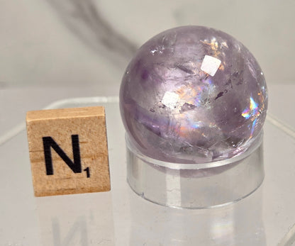 A faceted purple crystal sphere with iridescent internal reflections, displayed on a clear acrylic stand next to a wooden Scrabble tile showing the letter 'N'.