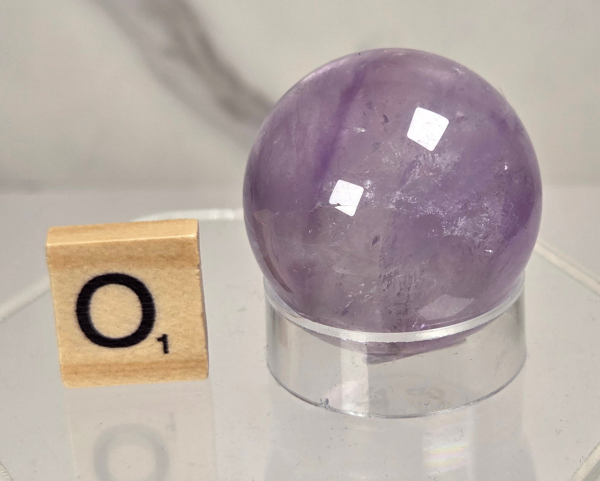 A polished purple amethyst crystal sphere on a clear acrylic stand, shown next to a wooden Scrabble tile with the letter O on a light marble surface.
