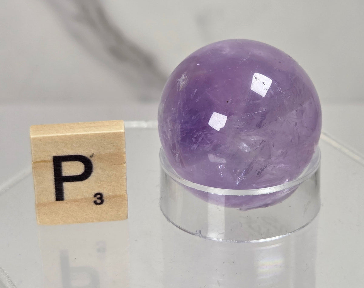 A polished purple amethyst crystal sphere resting on a clear acrylic display stand, next to a wooden Scrabble tile with the letter P, set against a soft-focus gray marble background.