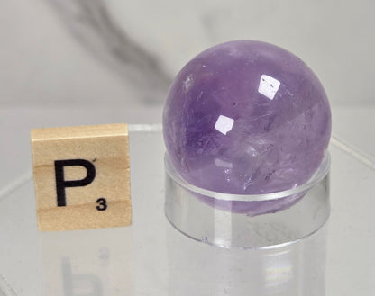 A polished purple amethyst crystal sphere resting on a clear acrylic display stand, next to a wooden Scrabble tile with the letter P, set against a soft-focus gray marble background.