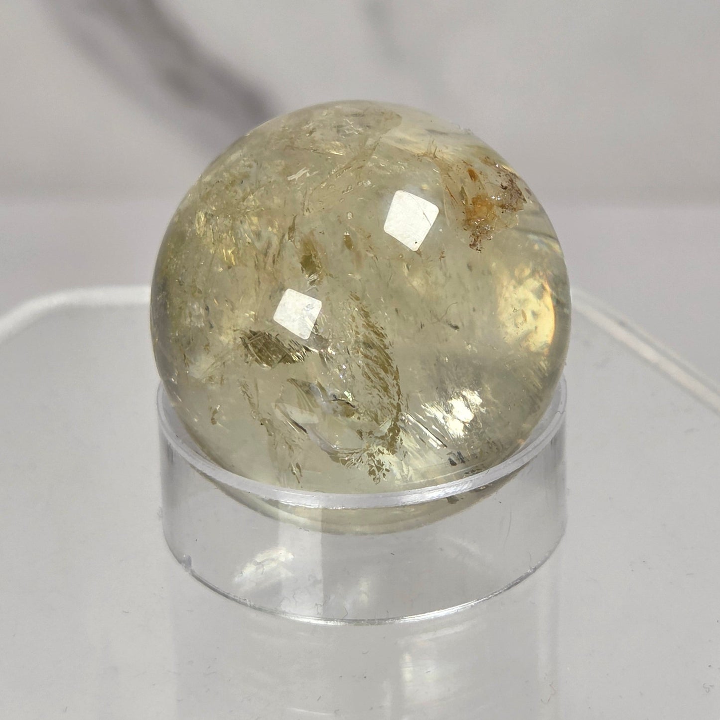A round, polished clear quartz crystal sphere with internal inclusions, displayed on a clear acrylic stand against a light gray marble-patterned background.