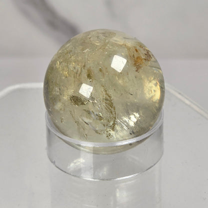 A round, polished clear quartz crystal sphere with internal inclusions, displayed on a clear acrylic stand against a light gray marble-patterned background.