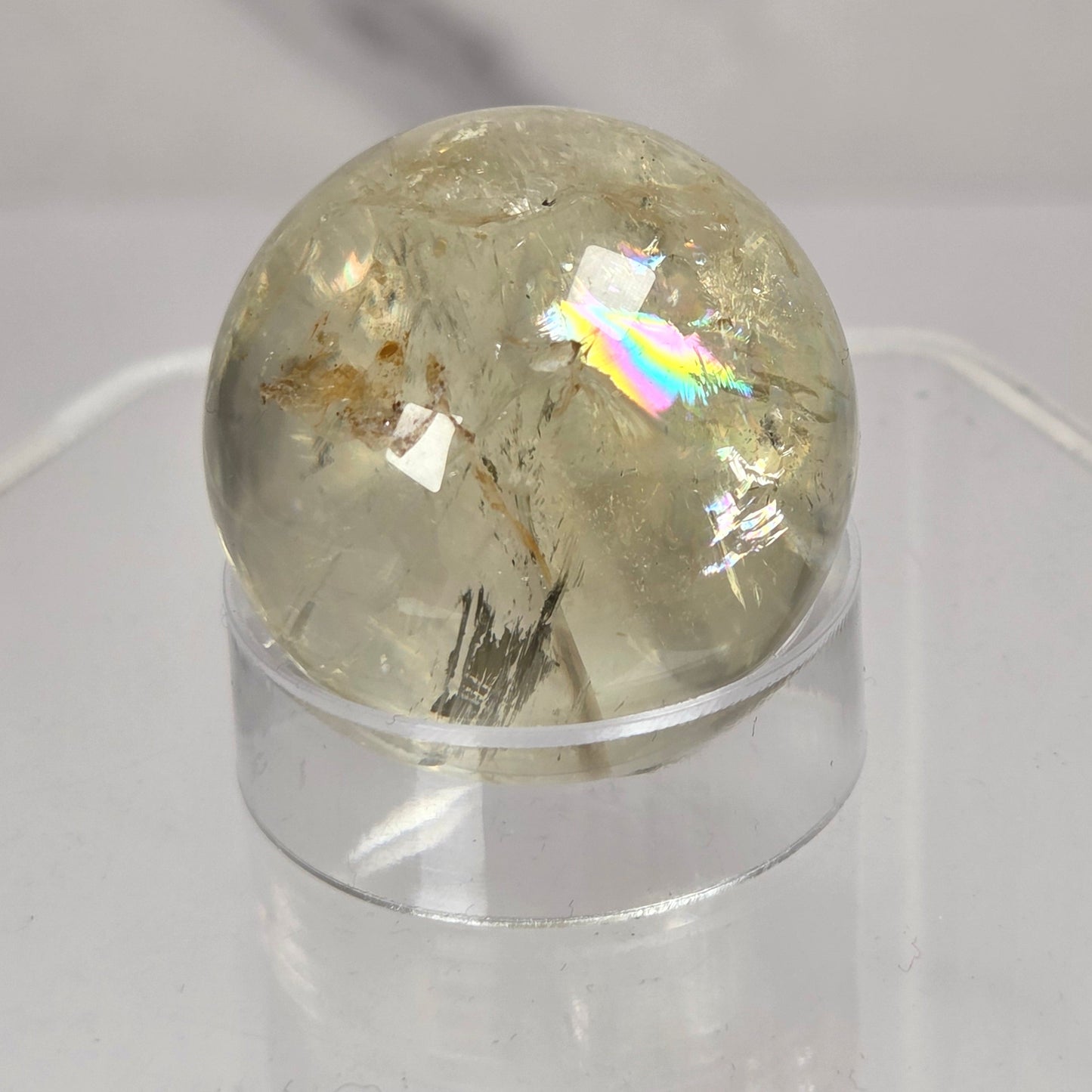 A faceted clear crystal sphere with internal inclusions and an iridescent rainbow sheen, displayed on a clear acrylic stand against a light gray background.