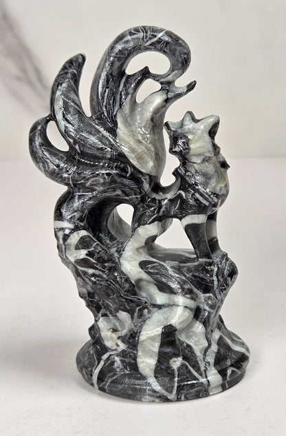 A small sculptural figurine of a fox-like creature with multiple curled tails, standing on a textured base. The object has a weathered, black and white coloring, set against a light marble-patterned background.