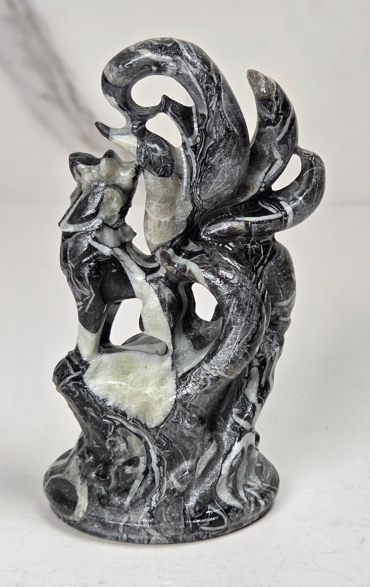 A small sculptural figurine of a fox-like creature with multiple curled tails, standing on a textured base. The object has a weathered, black and white coloring, set against a light marble-patterned background.