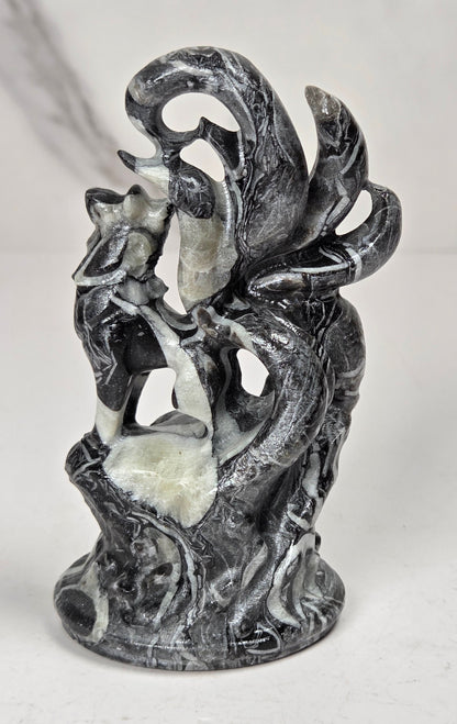 A small sculptural figurine of a fox-like creature with multiple curled tails, standing on a textured base. The object has a weathered, black and white coloring, set against a light marble-patterned background.