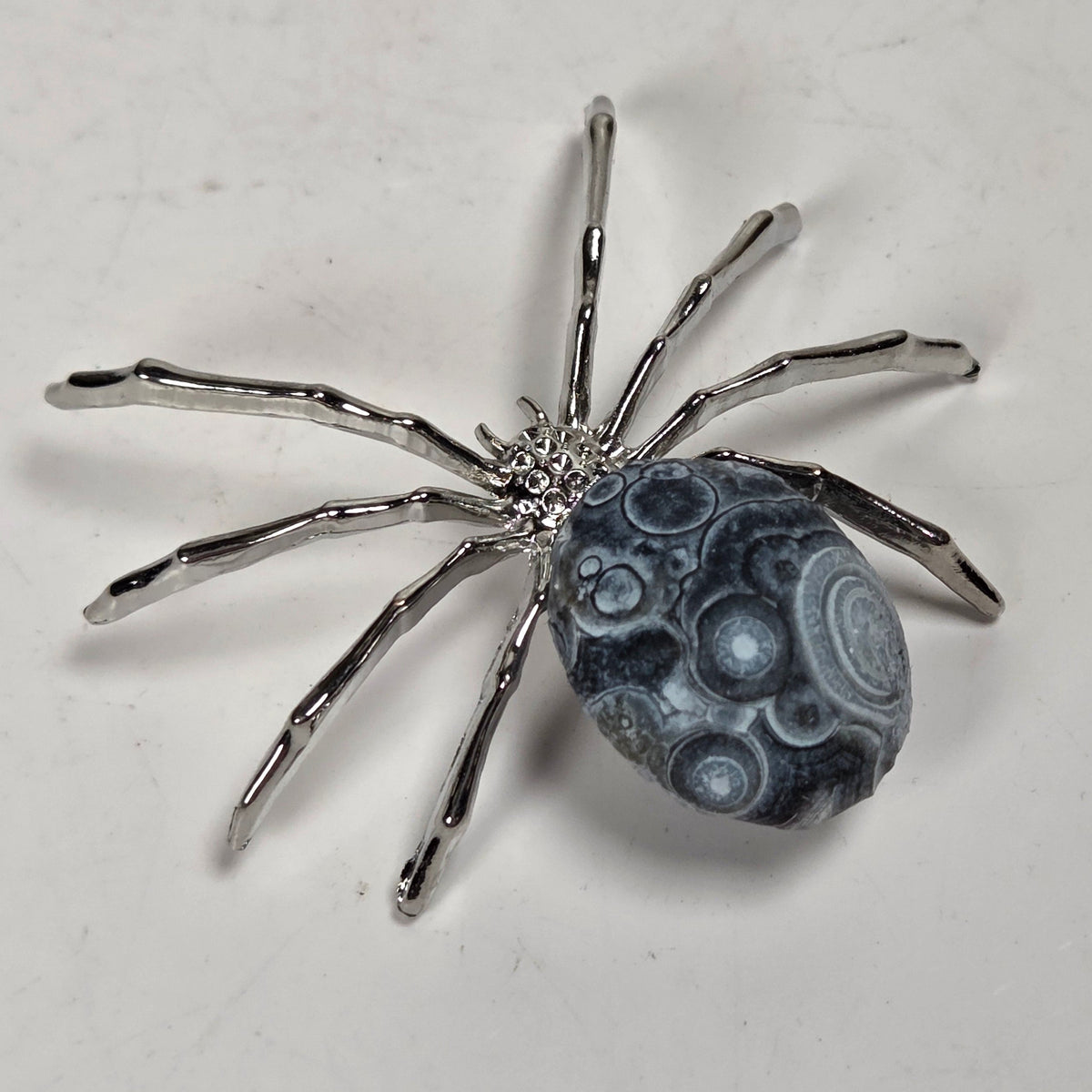 A spider-shaped pendant featuring a polished black bead body with white circular details and metallic silver legs, shown on a plain light background.