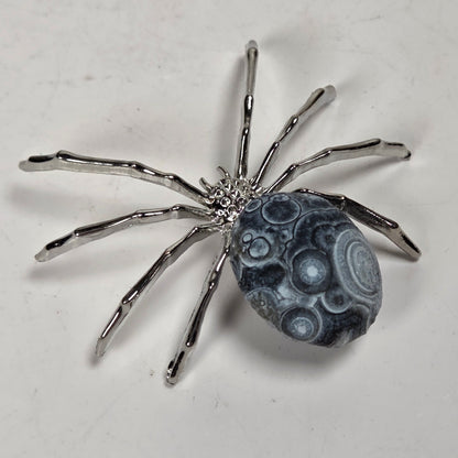 A spider-shaped pendant featuring a polished black bead body with white circular details and metallic silver legs, shown on a plain light background.