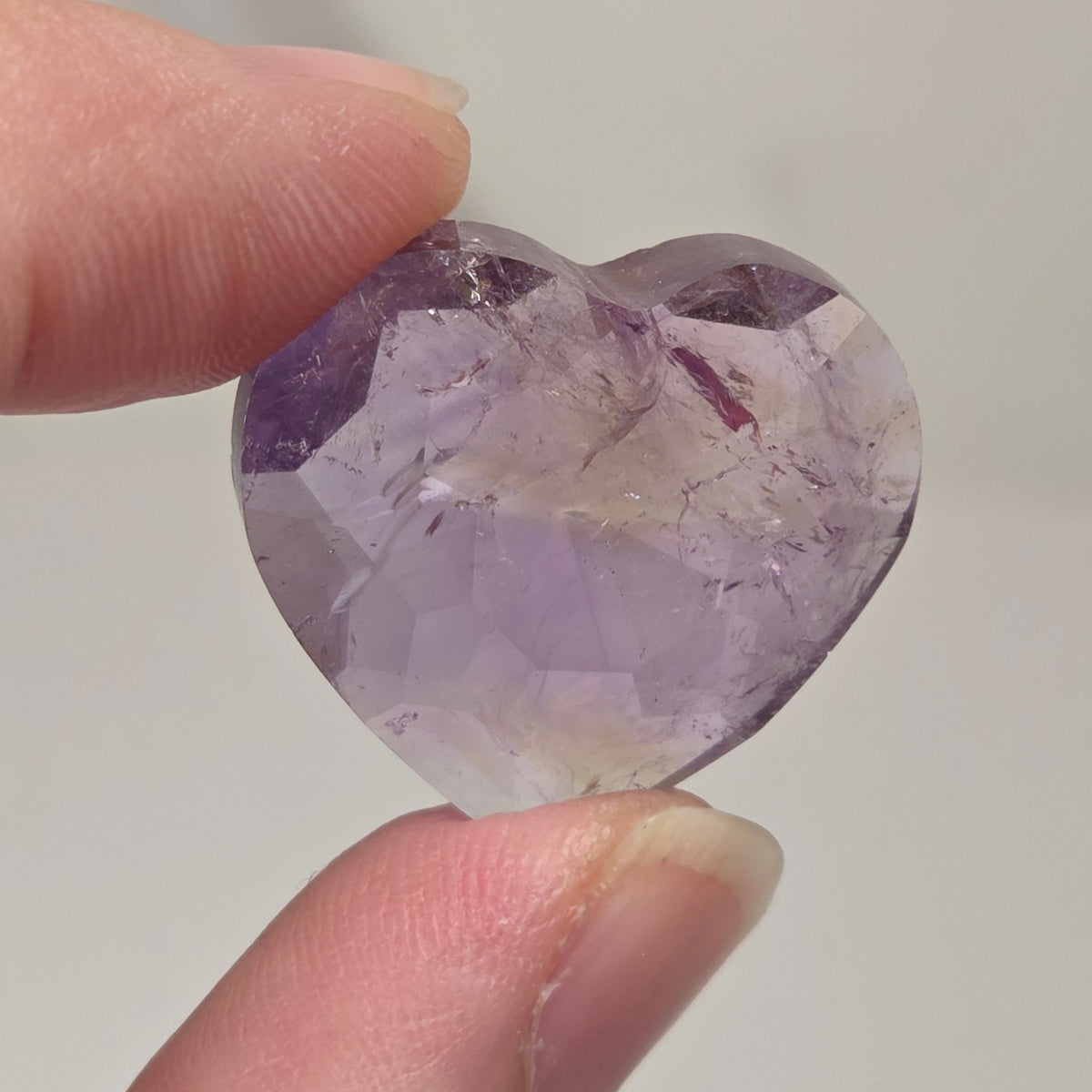 Faceted ametrine crystal heart being held between fingers against a white backrgound