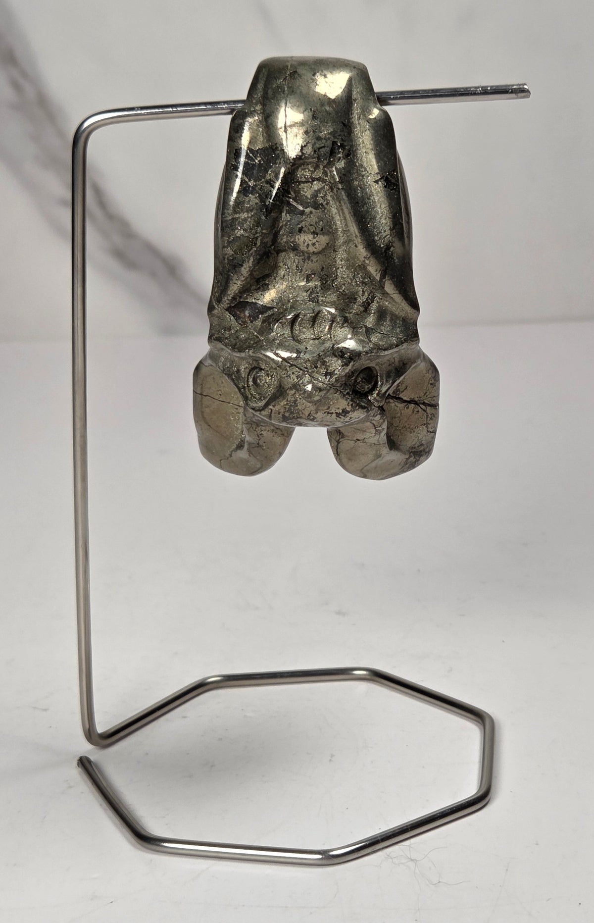 A pyrite sculptural bat carving a long, thin metal frame and a hexagonal loop, photographed against a white and light gray marble-textured background.
