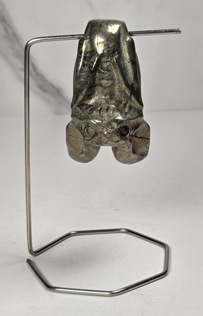 A pyrite sculptural bat carving a long, thin metal frame and a hexagonal loop, photographed against a white and light gray marble-textured background.