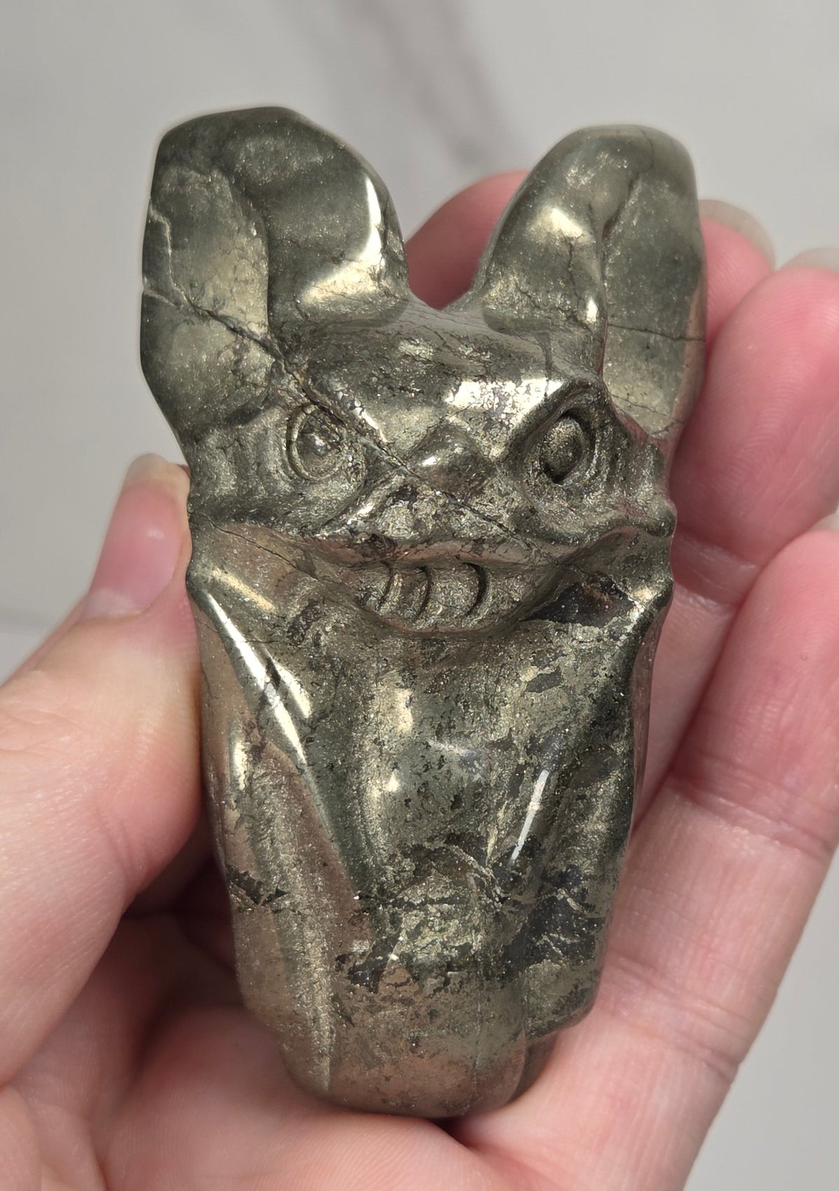 A pyrite sculptural bat carving being held in a hand, photographed against a white and light gray marble-textured background.