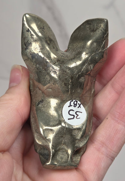 A pyrite sculptural bat carving being held in a hand, photographed against a white and light gray marble-textured background.