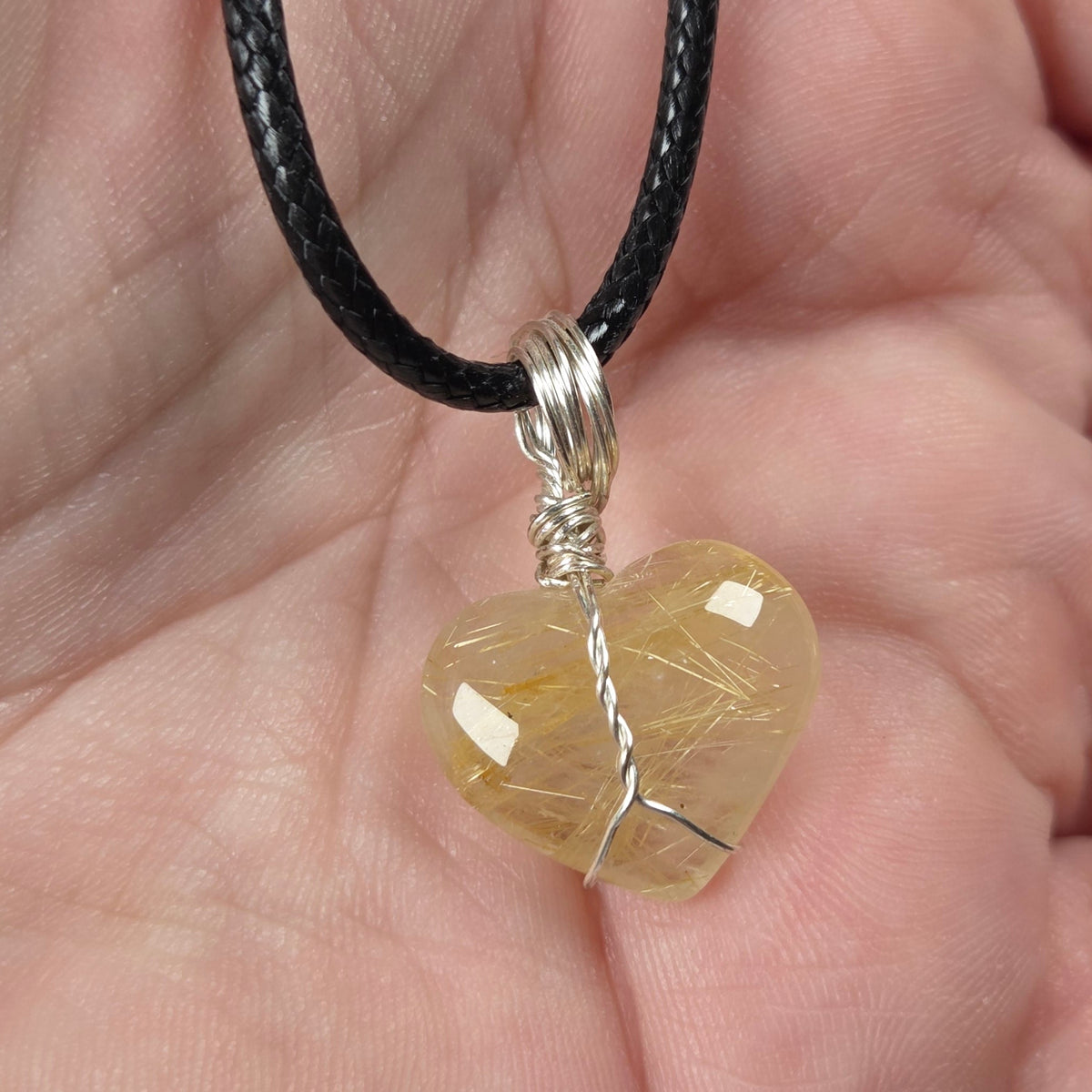 A heart-shaped pendant made of clear quartz stone wrapped in silver wire, hanging from a black cord and held in a person's hand.