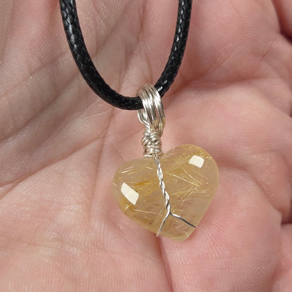 A heart-shaped pendant made of clear quartz stone wrapped in silver wire, hanging from a black cord and held in a person's hand.