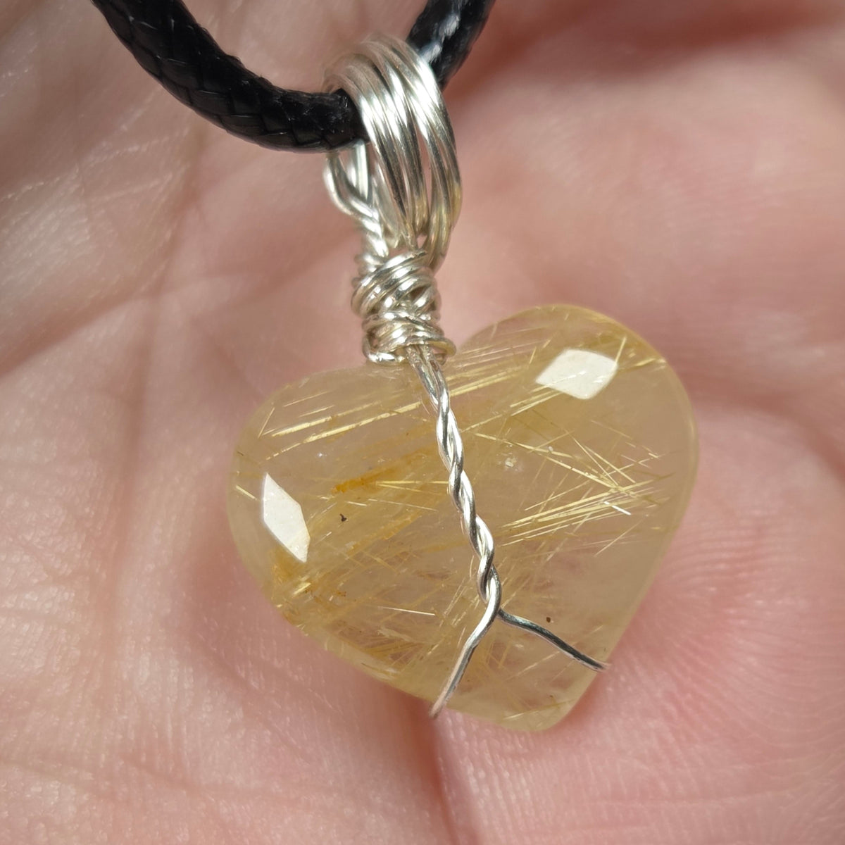 A heart-shaped pendant made of clear quartz stone wrapped in silver wire, hanging from a black cord and held in a person's hand.