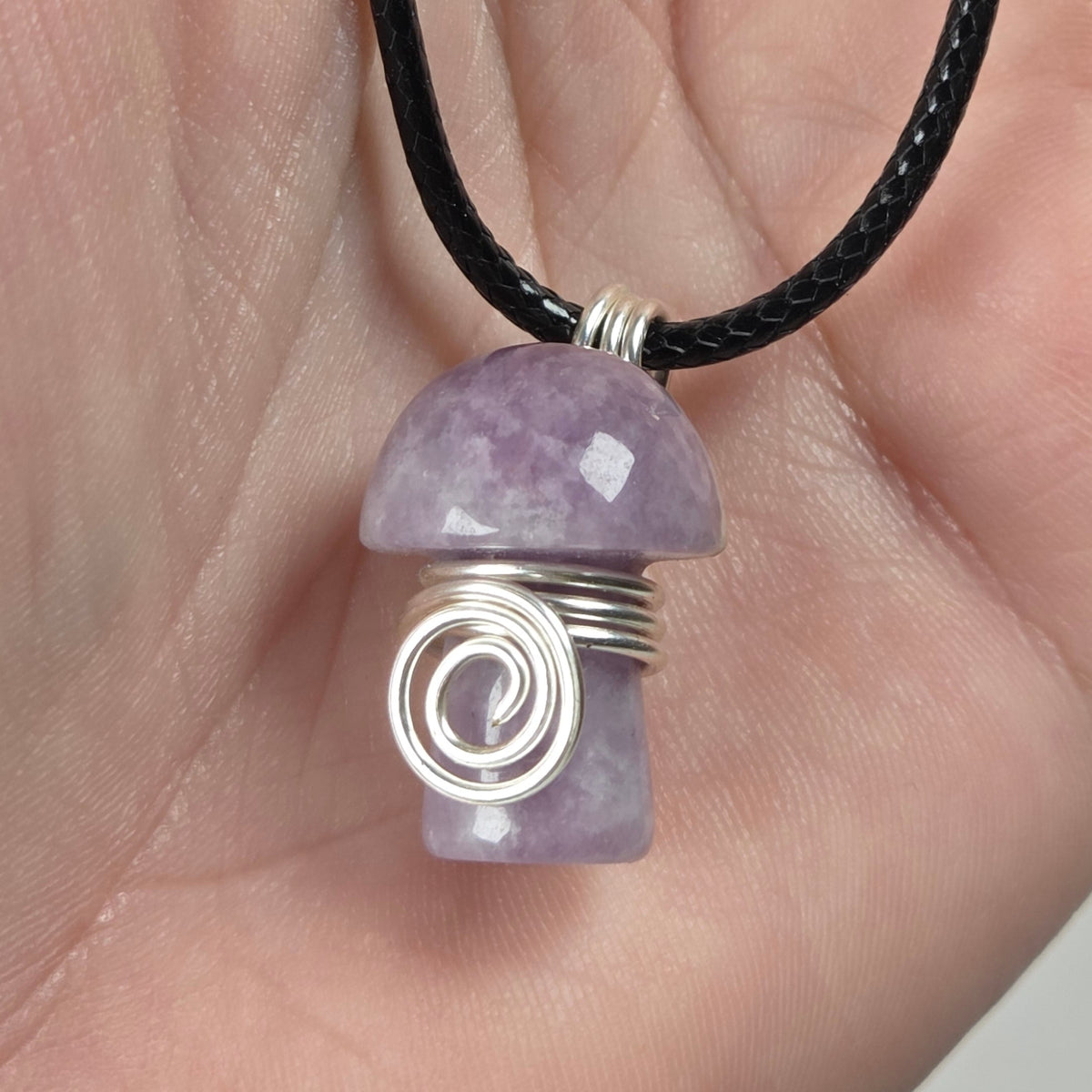 A mushroom-shaped pendant made of lepidolite stone wrapped in silver wire, hanging from a black cord and held in a person's hand.