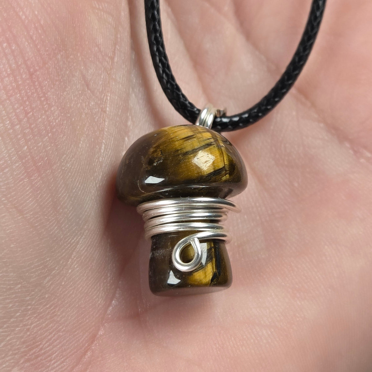 A mushroom-shaped pendant made of tigers eye stone wrapped in silver wire, hanging from a black cord and held in a person's hand.