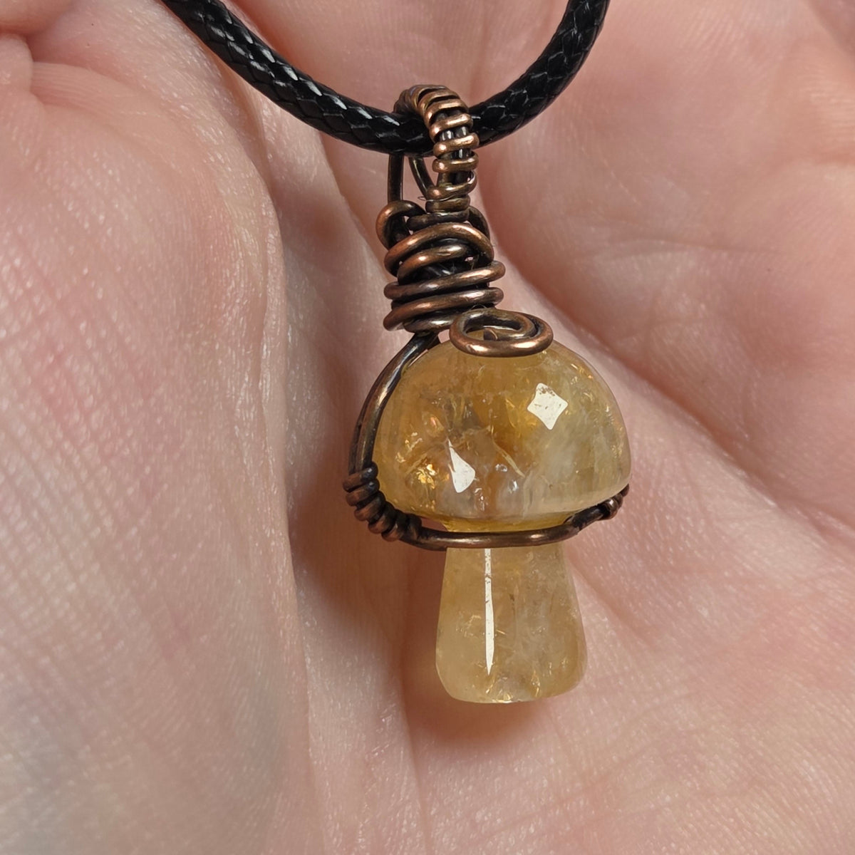 A mushroom-shaped pendant made of milky white stone wrapped in copper wire, hanging from a black cord and held in a person's hand.