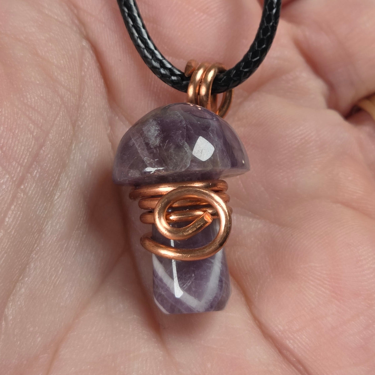 A mushroom-shaped pendant made of dream amethyst  wrapped in copper wire, hanging from a black cord and held in a person's hand.