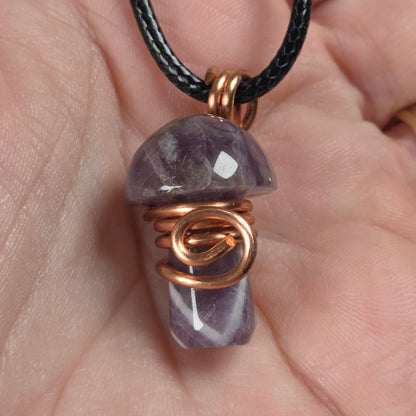 A mushroom-shaped pendant made of dream amethyst  wrapped in copper wire, hanging from a black cord and held in a person's hand.