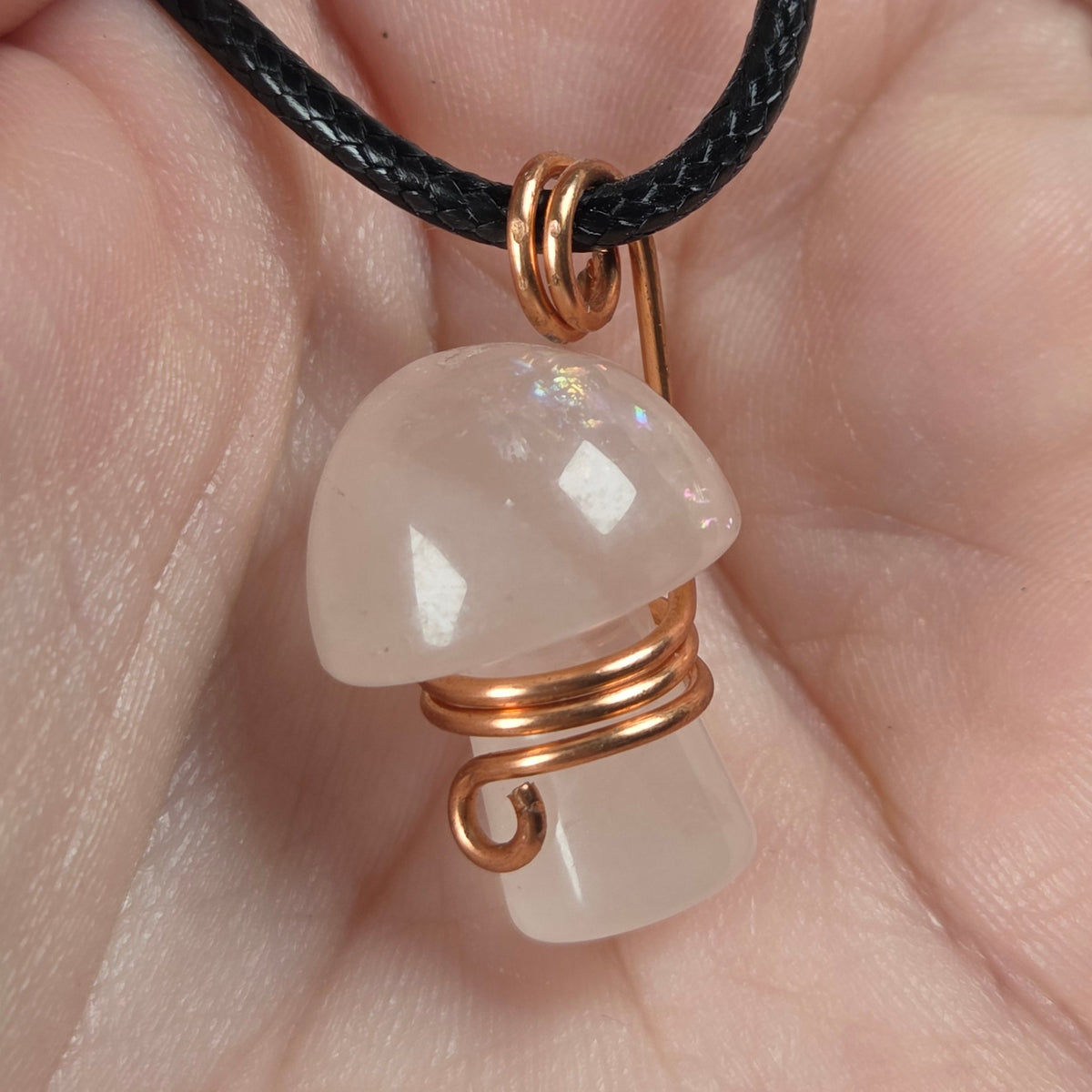 A mushroom-shaped pendant made of milky white stone wrapped in copper wire, hanging from a black cord and held in a person's hand.