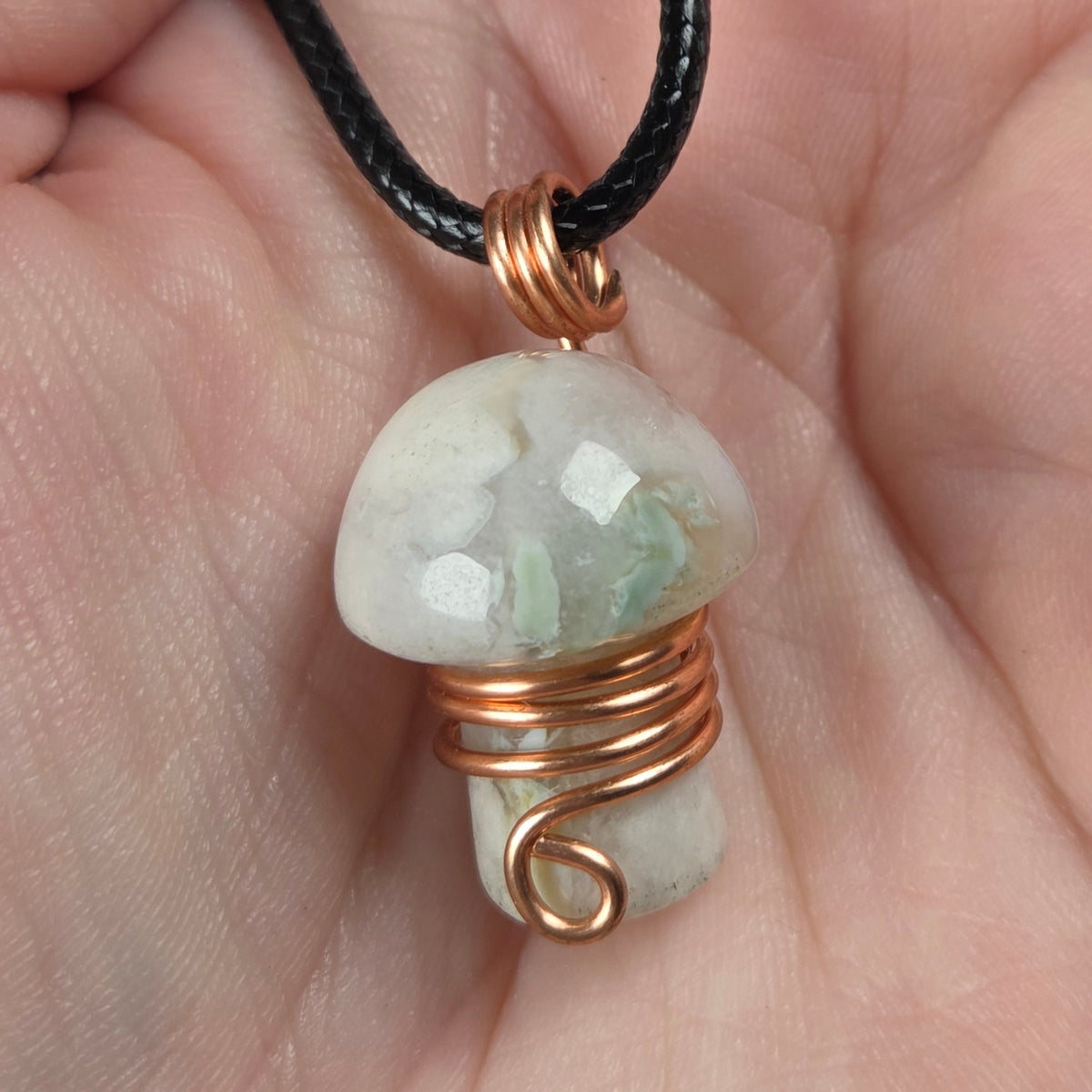 mushroom shaped crystal carving wrapped in copper wire with a black cord being held in a hand.