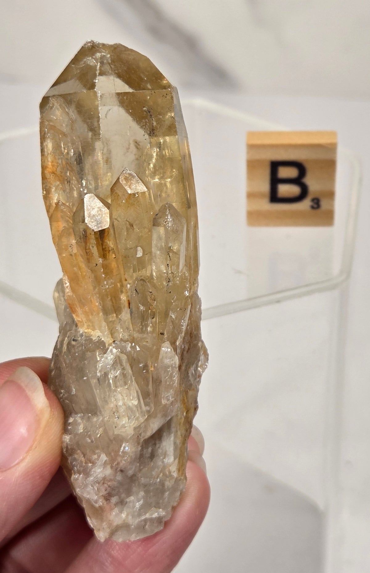 A hand holds a natural quartz crystal cluster with prominent golden-yellow and clear facets. A Scrabble tile with the letter 'B' is visible in the background on a white surface.