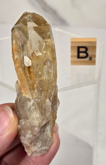 A hand holds a natural quartz crystal cluster with prominent golden-yellow and clear facets. A Scrabble tile with the letter 'B' is visible in the background on a white surface.